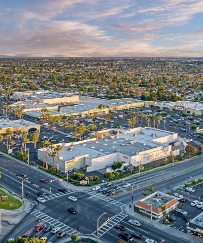 Aerial view of a suburban city at sunset with a large shopping center, busy roads, parking lots, and scattered palm trees.