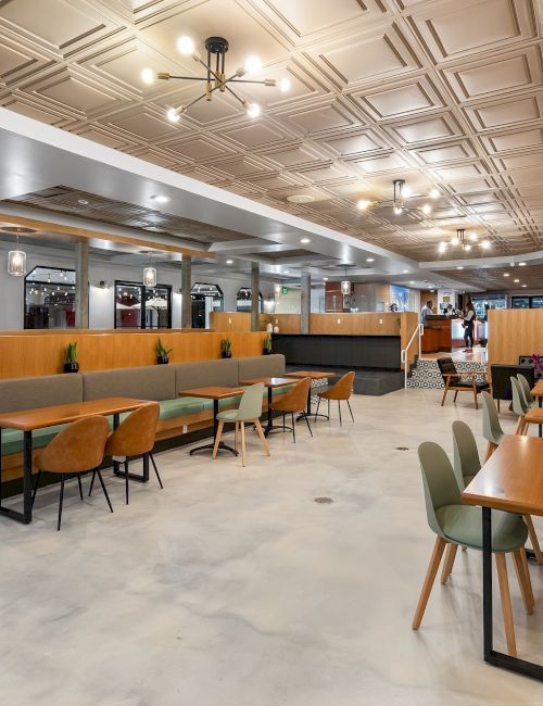 A bright modern lounge/cafeteria with mint booths, wooden tables, pendant lights, and patterned ceiling panels.