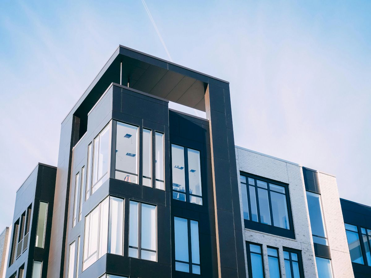 A modern building with dark glass and white walls rises against a blue sky, with contrails above.