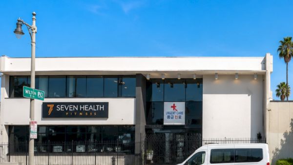 A building entrance with signs for Seven Health Fitness and Urgent Care, plus a parked van under a clear blue sky.